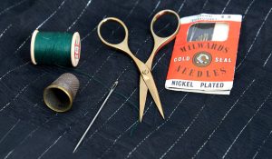 A picture of the gold plated embroidery scissors on pinstriped silk, with a vintage thimble, a pack of vintage sewing needles and green thread.